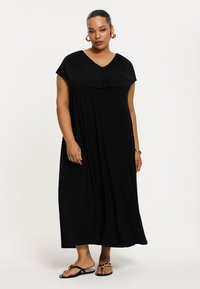 Robe maxi noire avec col plissé, manches courtes, design fluide et tissu lisse. Portée avec des sandales noires avec des accents dorés.