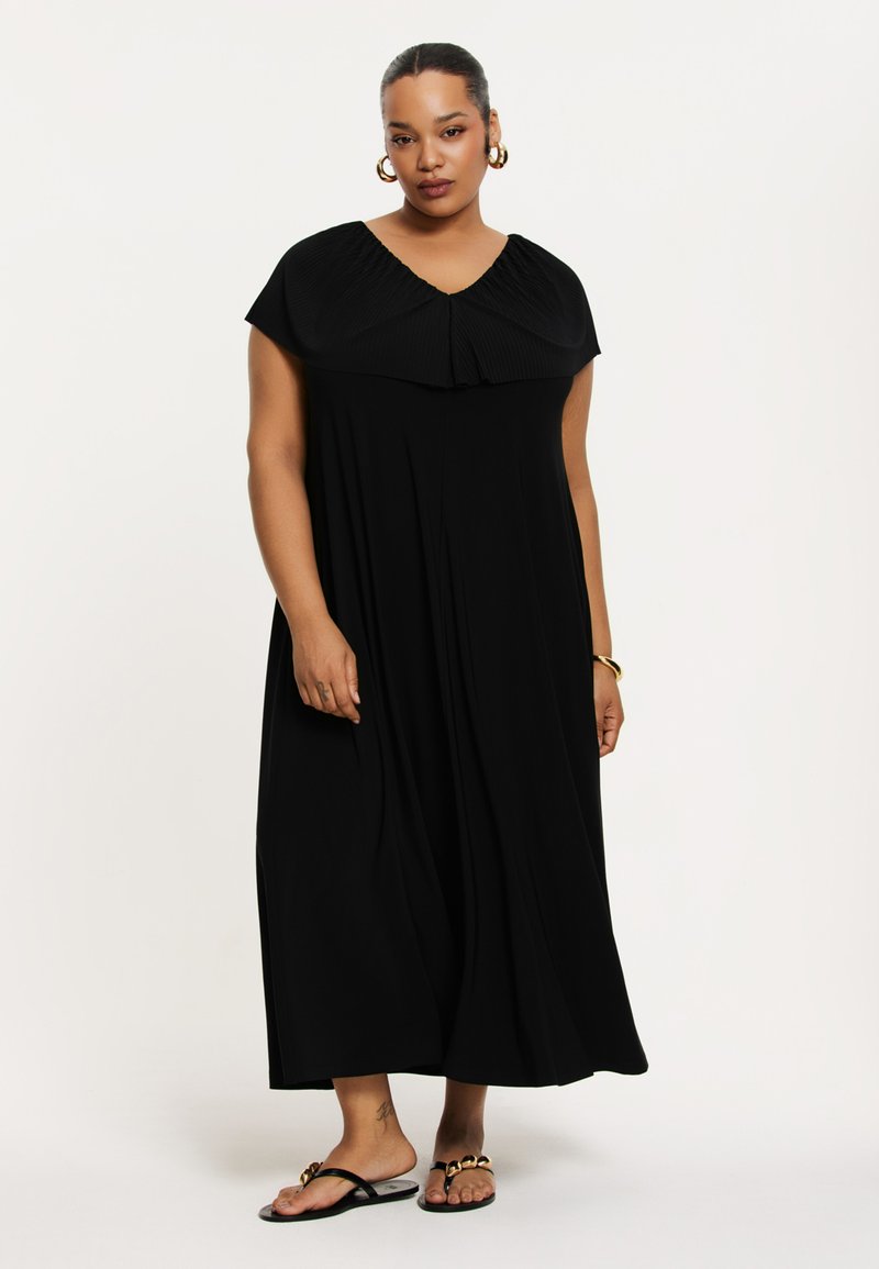 Robe maxi noire avec col plissé, manches courtes, design fluide et tissu lisse. Portée avec des sandales noires avec des accents dorés.