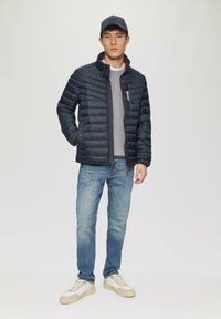 Námornícka pufrová bunda s horizontálnym prešívaním, skombinovaná so šedým sweatshirtom a svetlo modrými džínami. Obsahuje biele tenisky s béžovými akcentmi.