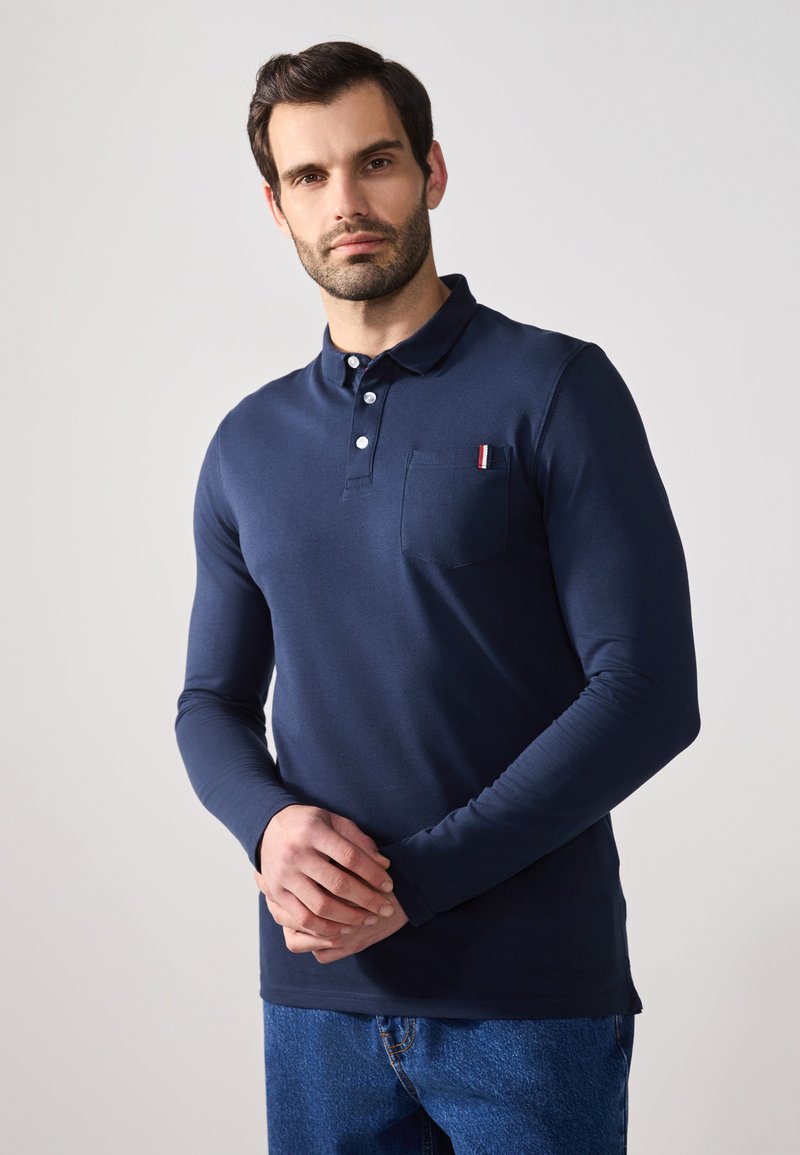 Polo de manga larga azul marino en mezcla de algodón, con un botón en el placket y un bolsillo en el pecho con acentos a rayas rojas y blancas.
