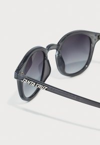 Schwarze und graue Sonnenbrille mit glänzender Oberfläche, runden Gläsern und kontrastierender "SANTA CRUZ"-Markierung an den Bügeln. Subtile Farbverlauf-Tönung der Gläser.