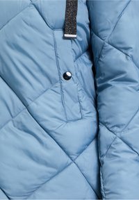 Jasnoniebieska kurtka puchowa z diamentowym pikowaniem, błyszczącym guzikiem, jedną kieszenią oraz czarnym teksturowanym paskiem jako akcentem. Miękka, gładka powierzchnia.
