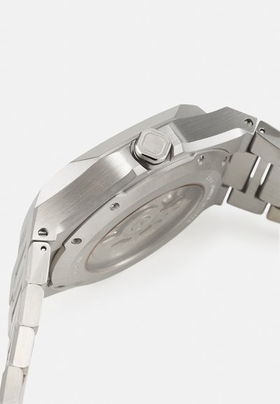 Srebrna ura iz nerjavečega jekla z osmokotno ohišjem, teksturirano površino in krožno prozorno zadnjo stranjo. Ima krono na strani.