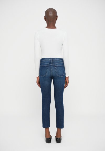 Persona che indossa una maglietta bianca a maniche lunghe, jeans blu scuro a vita alta con tasche posteriori e scarpe nere con borchie, in piedi di spalle su uno sfondo bianco.