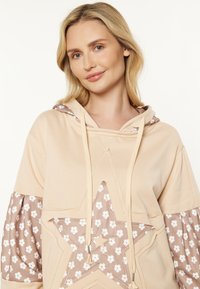 Beige hoodie med en stor stjärndesign, med ett blommönster på ärmarna. Mjuk material, dragsko och avslappnad passform.