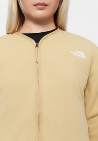 Veste zippée beige avec un col rond, fabriquée en tissu léger. Présente un logo brodé sur la poitrine gauche et des détails de zip visibles.
