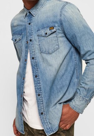 Mann trägt ein hellblaues Jeanshemd mit Knöpfen über einem weißen T-Shirt, eine Hand in der Tasche, vor neutralem Hintergrund.