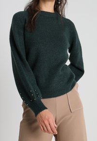 Vihreä neulepaita, jossa on pyöreä pääntie, röyhelöreuna ja teksturoitu kangas. Hihoissa on koristeellisia mustia helmiä. Yhdistetty vaaleanbeigeihin housuihin.