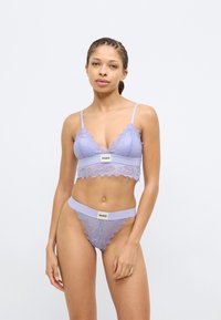 Modrček iz lavandinine čipke z nazobčanimi robovi in trakom z logotipom, združen s ujemajočimi se tangicami s podobnimi čipkastimi detajli in elastičnim pasom.