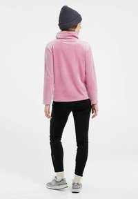 Pullover in velluto rosa con colletto alto, abbinato a leggings neri e sneakers grigie. Il pullover presenta maniche lunghe e una texture morbida.