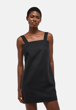 Femme aux cheveux courts portant une mini-robe noire sans manches avec de larges bretelles, debout devant un fond blanc uni.