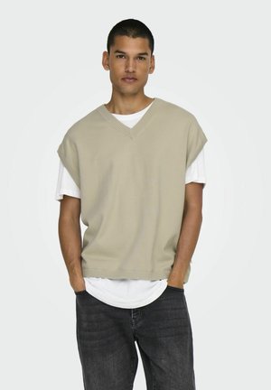 Beige gestrickter ärmelloser Pullover mit V-Ausschnitt, über einem weißen T-Shirt getragen, kombiniert mit dunklen Jeans und einem neutralen Hintergrund.