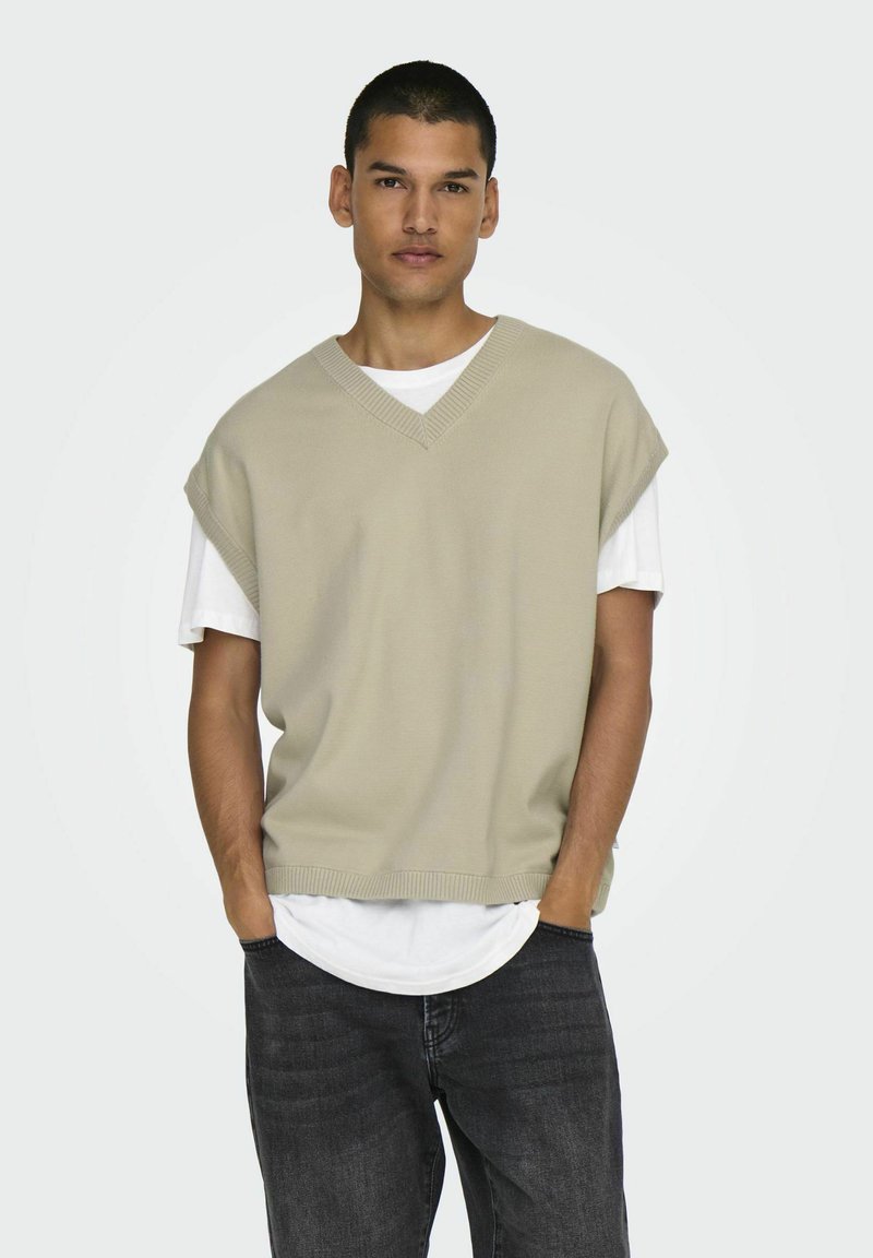 Beige gestrickter ärmelloser Pullover mit V-Ausschnitt, über einem weißen T-Shirt getragen, kombiniert mit dunklen Jeans und einem neutralen Hintergrund.