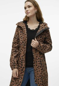 Brun mönstrad regnjacka med svarta leopardfläckar, justerbar dragsko i midjan, dragkedjestängning och huva. Modellen har en svart topp under.
