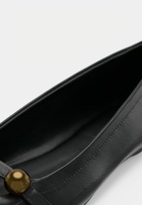 Μαύρο δερμάτινο παπούτσι με επίπεδη σόλα, που διαθέτει στρογγυλεμένη μύτη, λείο υφή, διακριτική ραφή και λεπτομέρεια με χρυσό κουμπί στο πάνω μέρος.