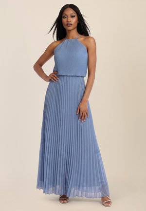 Woman with long straight hair wearing a sleeveless blue pleated halter-neck maxi dress and silver open-toe heels, standing with hand on hip.