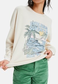 Valkoinen, tekstuurilla varustettu collegepaita, jossa on graafinen printti palmupuista, aalloista ja auringosta, kevyillä sinisen ja vaaleanpunaisen sävyillä.
