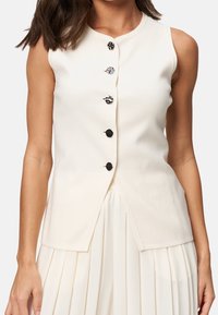 Femme portant un gilet blanc sans manches à cinq boutons décoratifs sombres, associé à une jupe plissée blanche.