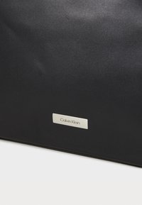 Svart laptopveske med jevn tekstur, rektangulær form og sølvfarget Calvin Klein-merket metalltag foran.