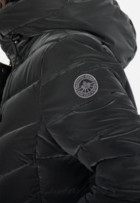 Doudoune noire avec une finition brillante, dotée d'une capuche amovible, d'un design matelassé et d'un patch avec logo sur le haut du bras.
