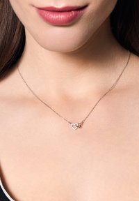 Srebrny naszyjnik z łańcuchem, złożony z dwóch splecionych zawieszek w kształcie serc, jedno o błyszczącej fakturze, a drugie gładkie, oba na nagiej szyi.