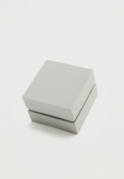 Κλειστό ανοιχτό γκρι τετράγωνο κουτί με διακριτικό ανάγλυφο λογότυπο στην κορυφή, τοποθετημένο σε λευκό απλό φόντο.