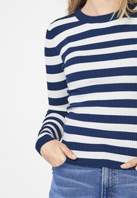 Námořnický modrý a bílý pruhovaný pletený svetr s žebrovanou texturou, kulatým výstřihem a dlouhými rukávy, nošený se založenýma rukama v kapsách džín.