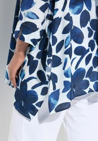 Blusa floreale con audaci motivi a foglia blu su uno sfondo chiaro, realizzata in un tessuto morbido. Le fessure laterali migliorano il flusso d'aria e il movimento.