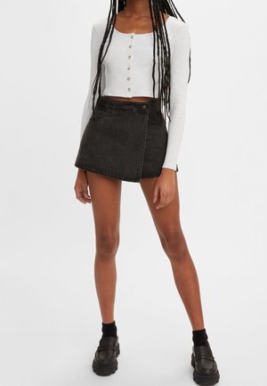 Frau trägt ein weißes, langärmeliges, geknöpftes Crop-Top, einen schwarzen Wickel-Minirock, schwarze Socken und schwarze klobige Schuhe.