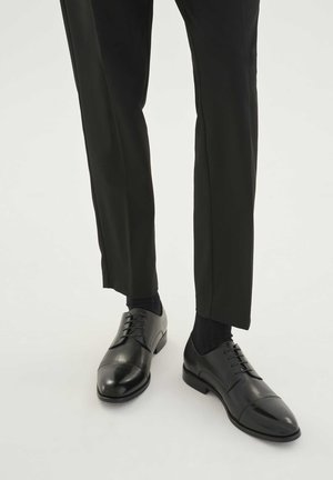 Černé kožené společenské boty s hladkým povrchem, zaoblenou špičkou a šněrováním; spárované s tmavými ponožkami a upravenými černými kalhotami.