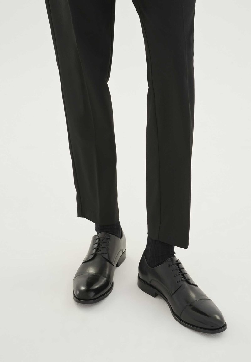 Chaussures habillées en cuir noir avec une finition lisse, bout arrondi et design à lacets ; associées à des chaussettes foncées et un pantalon noir ajusté.