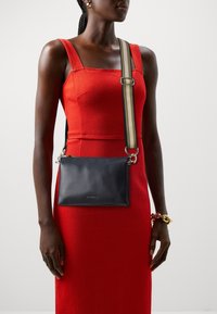 Donna che indossa un abito rosso aderente e orecchini a cerchio dorati, mentre porta una borsa a tracolla nera con una tracolla beige e nera.