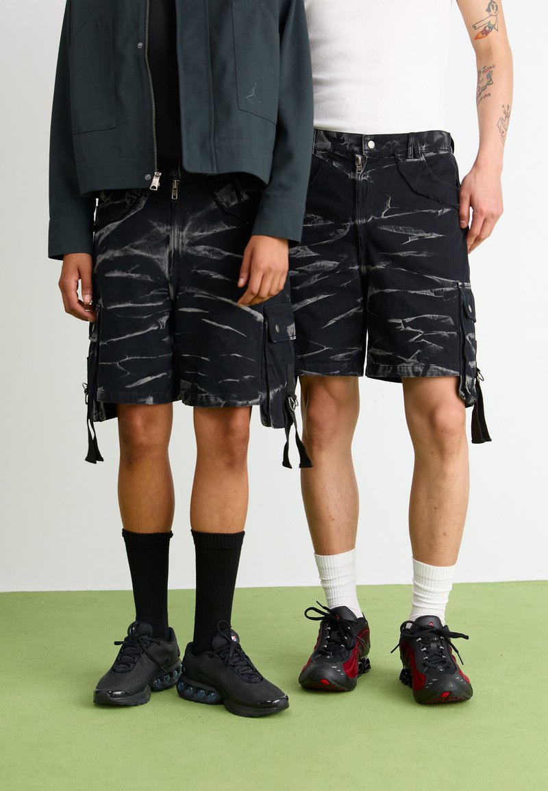 Schwarze Cargo-Shorts mit einem verwaschenen Design und weißen Streifen, zwei seitlichen Taschen und verstellbaren Kordeln, kombiniert mit sportlichen Sneakern.