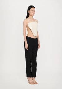 Top asimetric bej cu bretele subțiri, asortat cu pantaloni negri din stofă. Modelul stă în profil, evidențiind silueta și texturile distincte.