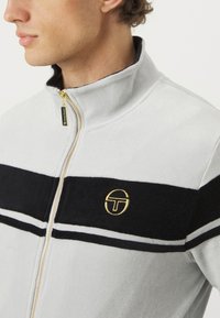 Sammenløft jakke i lys grå fleece med svarte og hvite striper. Har en gulllogo på brystet og gull glidelås-detaljer.