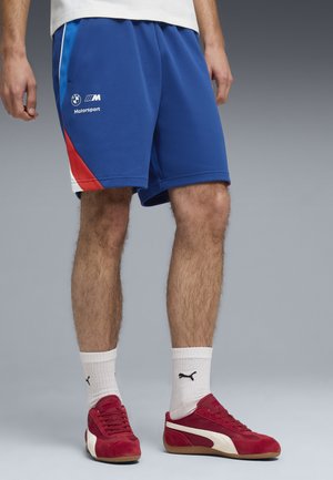 Mann trägt blaue BMW Motorsport-Shorts, weiße Puma-Socken und rote Puma-Sneaker, steht vor einem grauen Hintergrund.