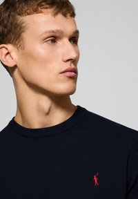 Marinblå tröja med rund hals, ribbad krage och en liten röd broderad logotyp på bröstet. Slät stickad textur, minimalistisk design.