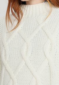 Sweter w kolorze kremowym z splotem w warkocze, z wysokim dekoltem i fakturowanymi wzorami, o luźnym kroju. Miękki, gruby materiał dzianinowy.
