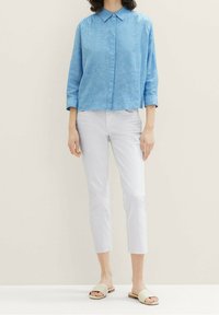 Chemise boutonnée en lin bleu clair avec un col, portée avec un pantalon blanc coupé au-dessus de la cheville et des sandales beige à enfiler. Texture lisse.