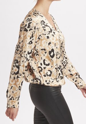 Frau trägt eine beige-schwarze Langarmbluse mit Leopardenmuster, kombiniert mit schwarzen, eng anliegenden Hosen, seitlich vor einem schlichten Hintergrund posierend.