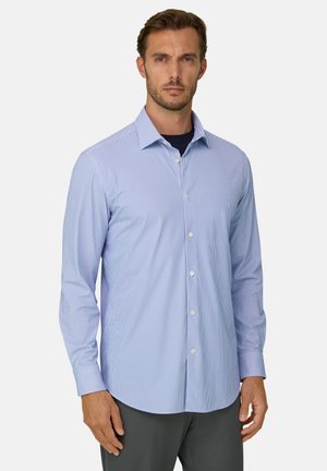 Homme portant une chemise bleu clair à fines rayures à manches longues avec boutons, et un pantalon sombre, sur un fond clair uni.