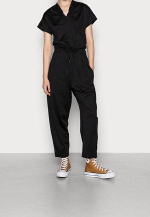 Combinaison noire à manches courtes, avec taille à cordon de serrage et deux poches avant. Portée avec des baskets montantes marron et semelles blanches.
