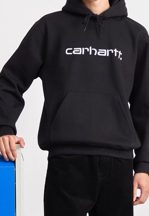 Osoba w czarnej bluzie z kapturem Carhartt z białym nadrukiem logo, jedna ręka spoczywa na niebiesko-białym przedmiocie, widoczne czarne spodnie.