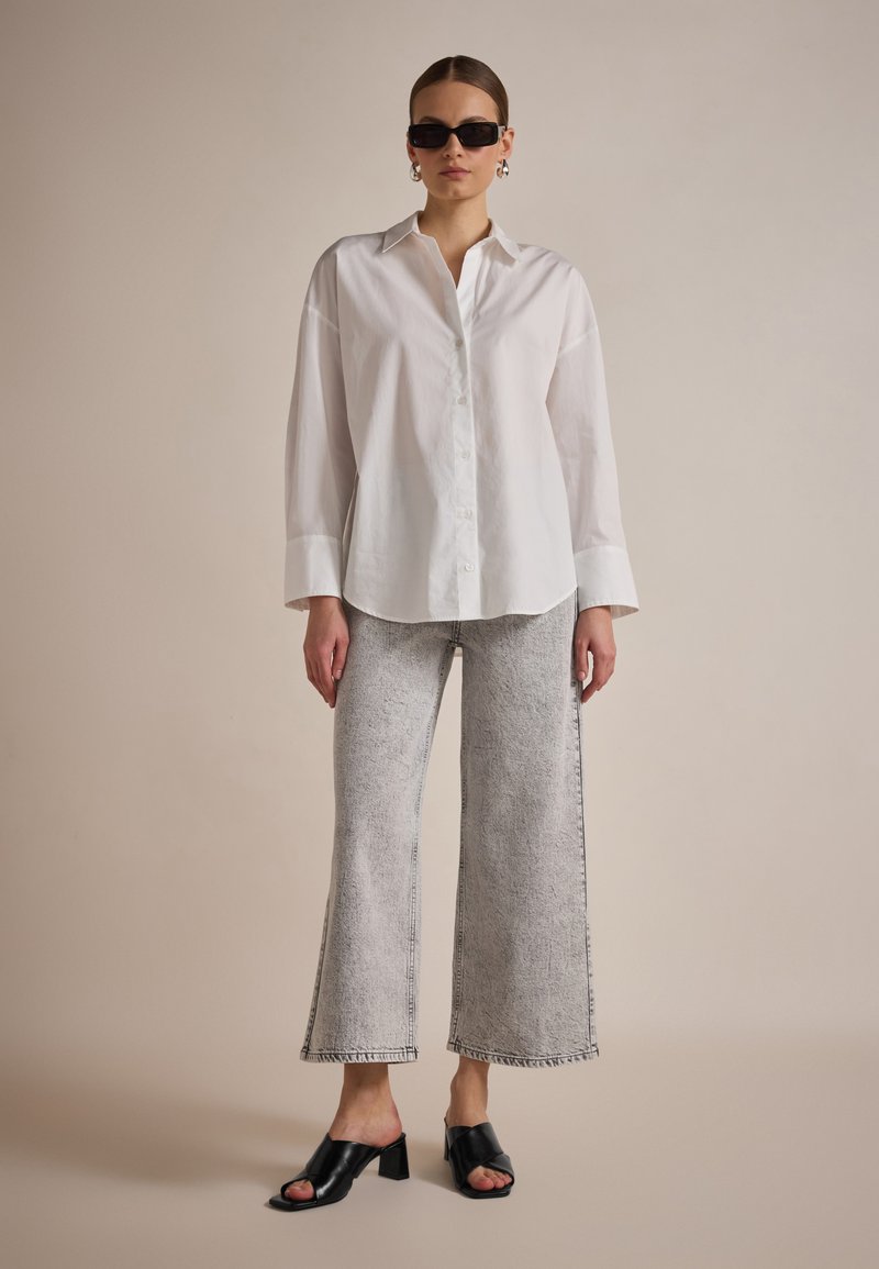 Camicia bianca con bottoni, pantaloni ampi in denim grigio chiaro, mule nere con tacco, e occhiali da sole neri a forma quadrata. Tessuto liscio e orli sfrangiati.