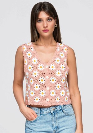 Ung kvinde iført ærmeløs lyserød hæklede top med hvide og gule marguerit mønstre og lyseblå denimjeans, hånd i lommen, neutral baggrund.