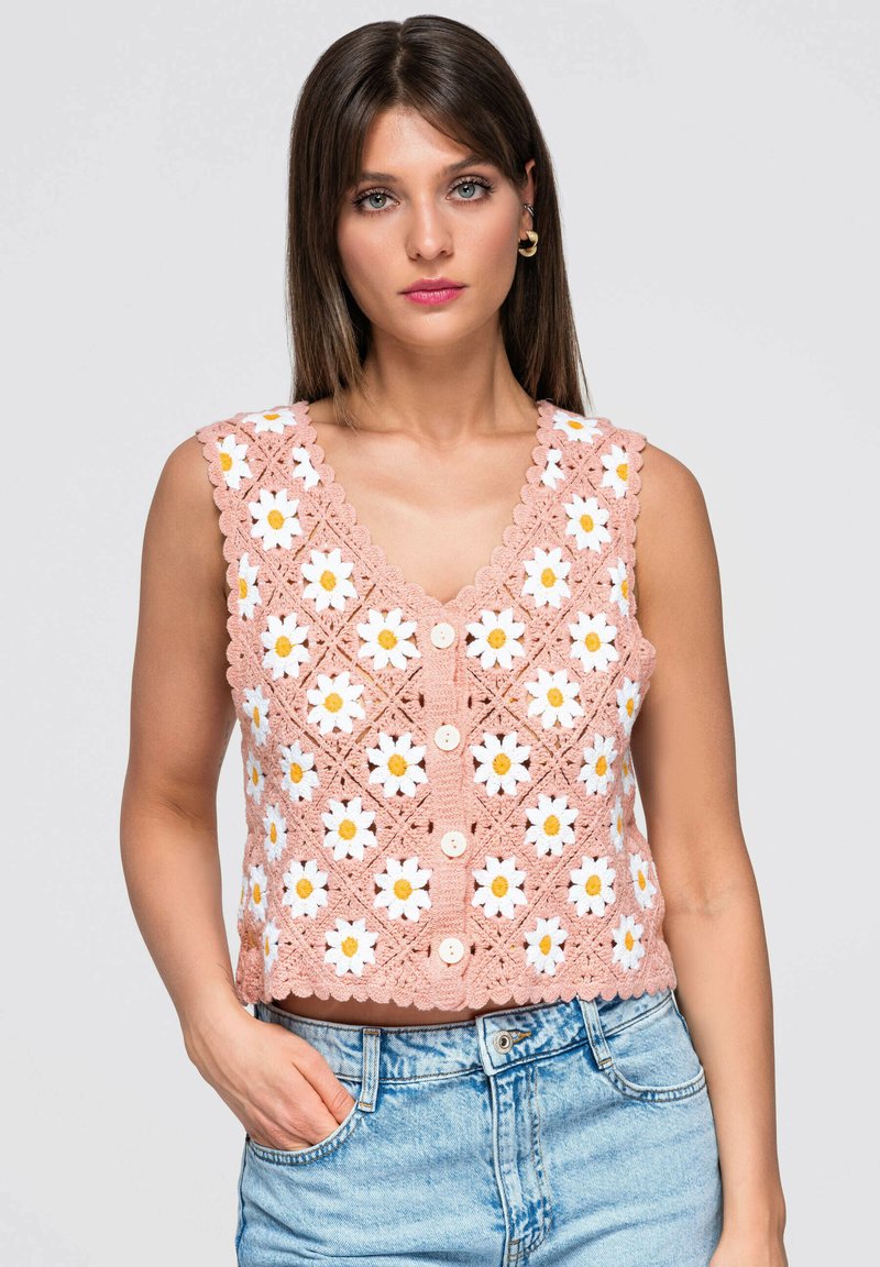 Jeune femme portant un haut crochet sans manches rose avec des motifs de marguerites blanches et jaunes et un jean en denim bleu clair, main dans la poche, fond neutre.