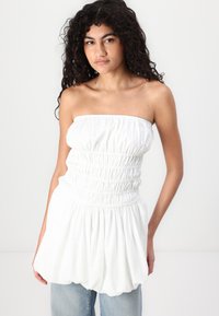 Λευκή αμάνικη μπλούζα φτιαγμένη από μαζεμένο ύφασμα, με εφαρμοστό κορσέ και φαρδύ κάτω στρίφωμα. Λείος υφασμάτινος χαρακτήρας, χωρίς σχέδια ή διακοσμήσεις.