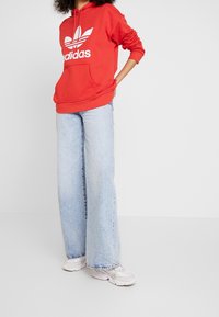 Person i röd Adidas-hoodie, ljusblå vida jeans och vita sneakers som står mot en enkel ljus bakgrund med händerna bakom ryggen.