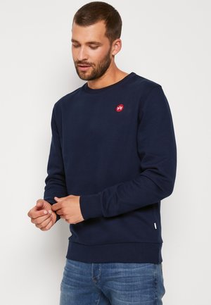 Granatowy sweter wykonany z miękkiego materiału, z okrągłym dekoltem, ściągaczami przy mankietach oraz okrągłym czerwonym logo na klatce piersiowej.