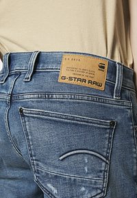 Šviesiai mėlynos džinsinės kelnės su rusvos odos G-Star Raw etikete ant juosmens, su klasikinio penkių kišenių dizainu ir matomomis siūlėmis.